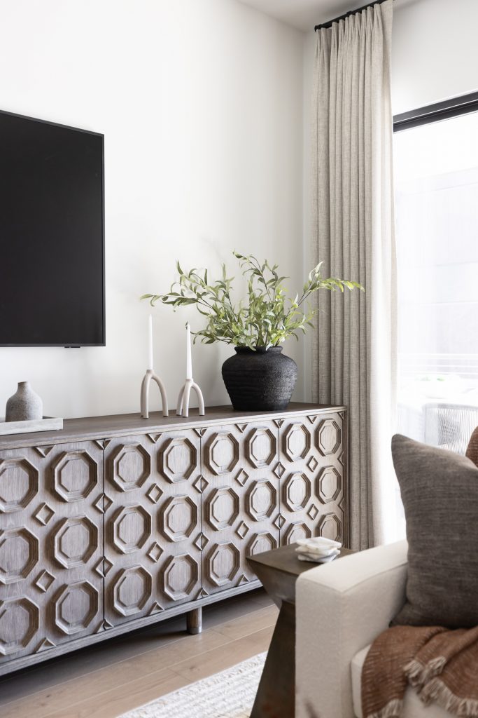 A wooden sideboard with geometric patterns holds two white candlesticks, a black vase with green foliage, and a small gray vase—Living with Lolo.