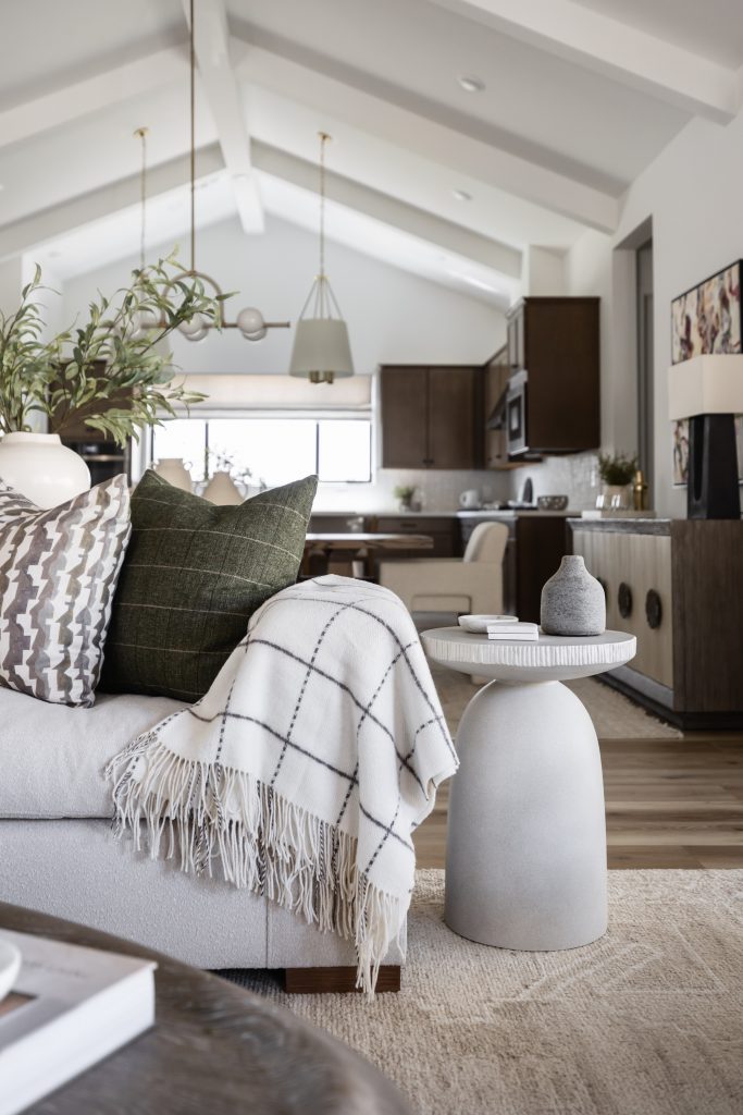 Modern living room with neutral furniture, patterned cushions, a plaid throw, round side table, and kitchen in the background—Living with Lolo.
