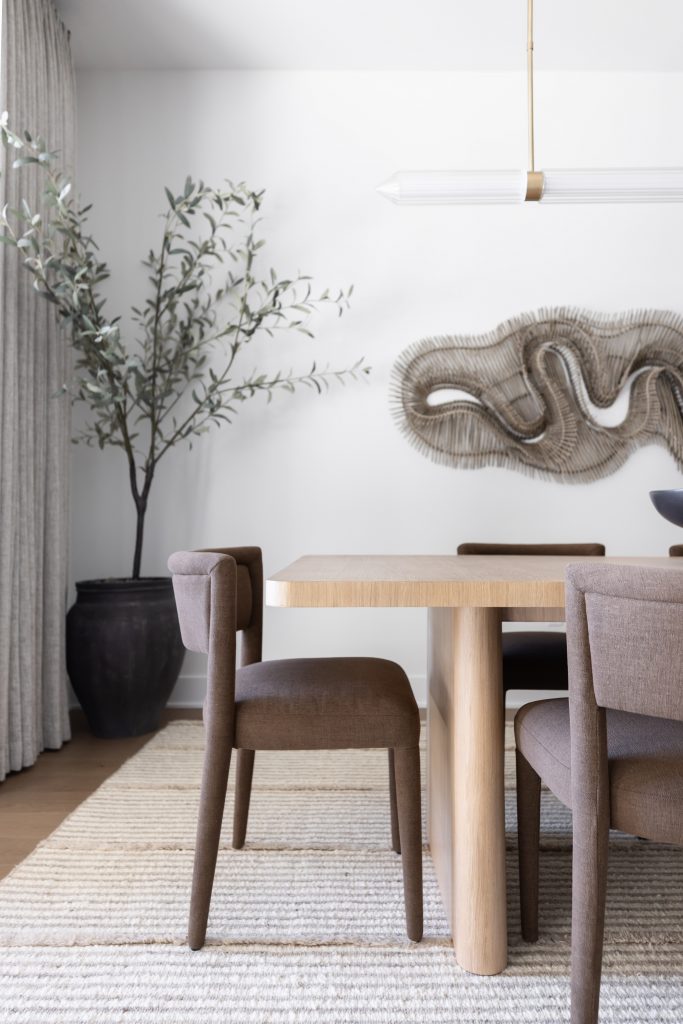 A modern dining room with a light wood table, brown upholstered chairs, a potted plant, abstract wall art, and tall curtains. — Living with Lolo.