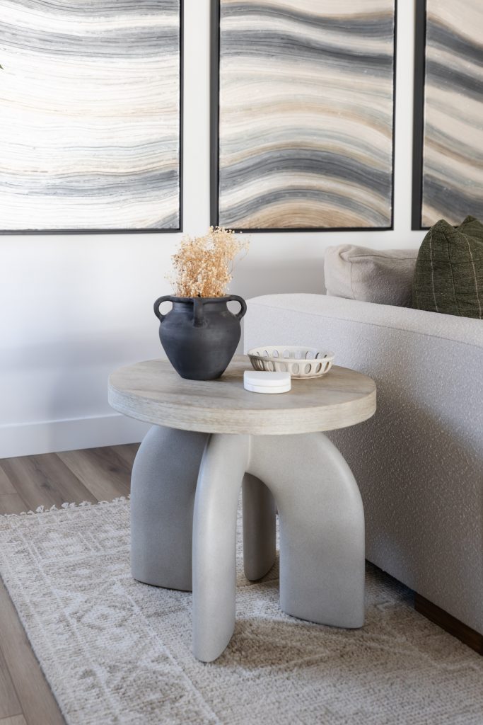 A modern side table with a black vase of dried flowers, a small dish, and decor stands by a light sofa and abstract wall art—Living with Lolo.