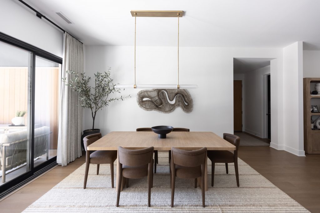 Modern dining room with a wooden table, six brown chairs, black bowl centerpiece, wall art, floor plant, and large curtained window.— Living with Lolo.