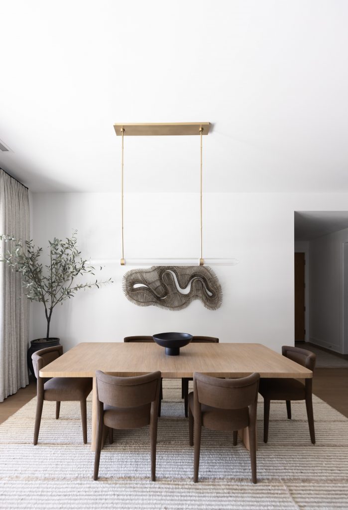 A modern dining room with a wooden table, six brown chairs, a black bowl, wall sculpture, potted plant, and ceiling light fixture—Living with Lolo.