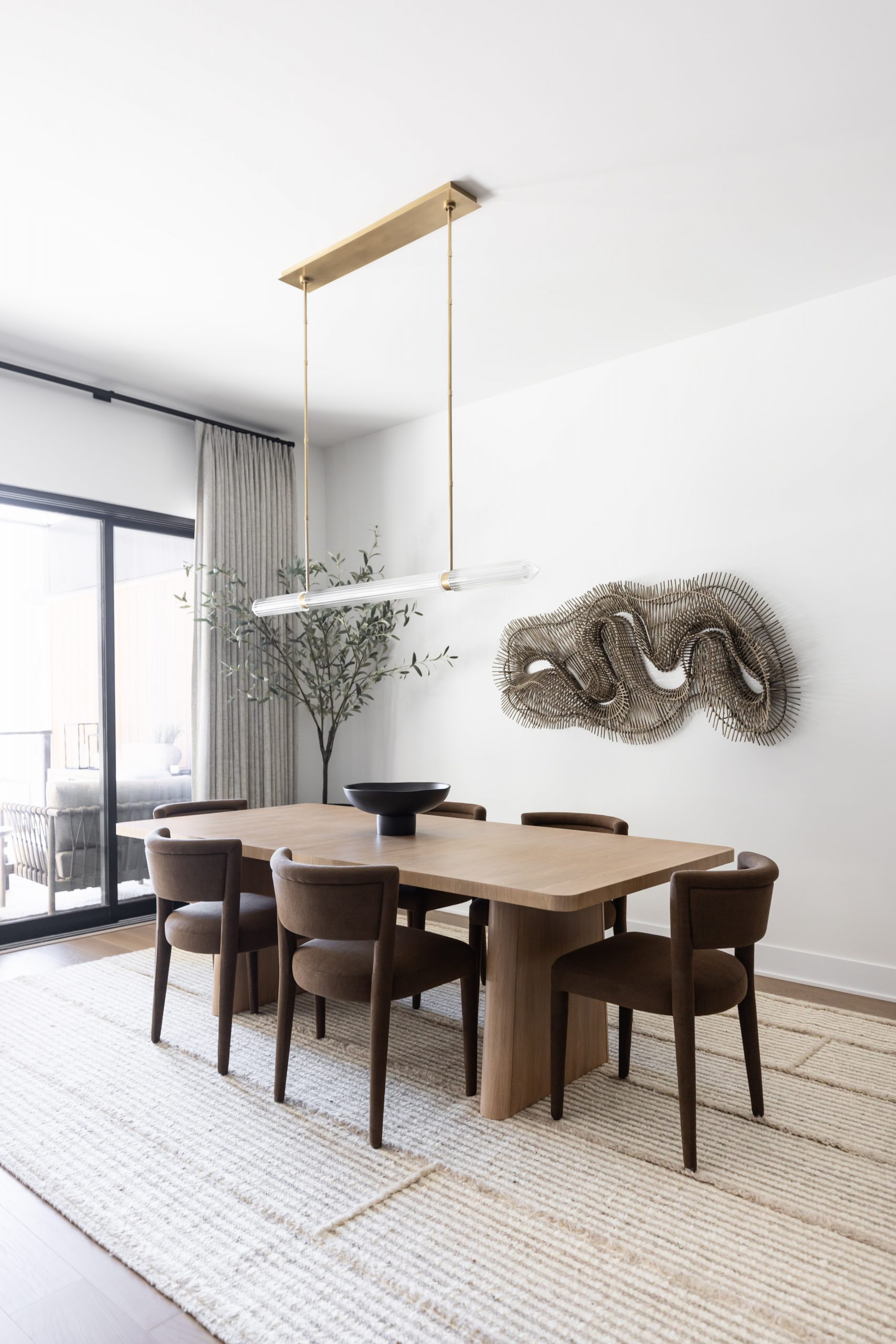 A dining room with a wooden table, six brown chairs, black bowl centerpiece, large window, wall sculpture, and hanging light—Living with Lolo.