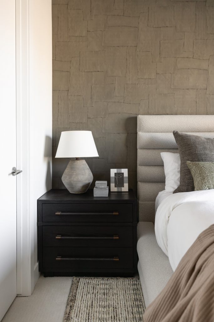 A modern bedroom includes a dark nightstand with a lamp, books, framed photo, neutral bedding, and a textured accent wall— Living with Lolo.