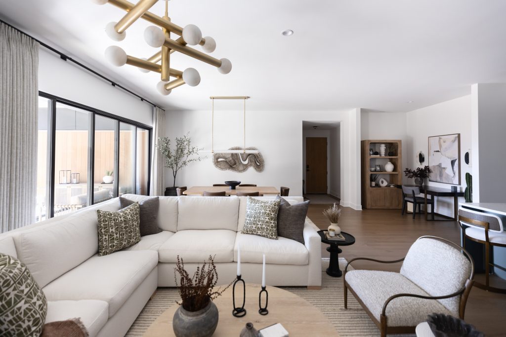 Modern living room with a white sectional sofa, accent chairs, round coffee table, large windows, and contemporary light fixtures. Dining area and decorative wall art in the background—Living with Lolo.