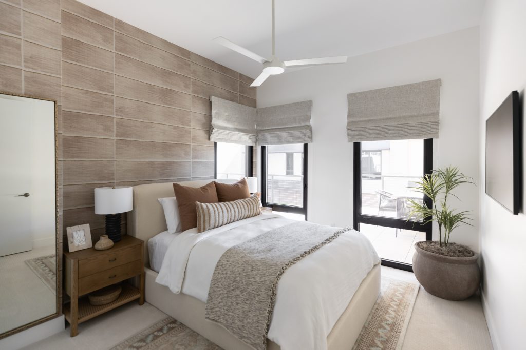 Modern bedroom featuring a bed with neutral bedding, wooden nightstand, large mirror, potted plant, TV, and two windows—Living with Lolo.