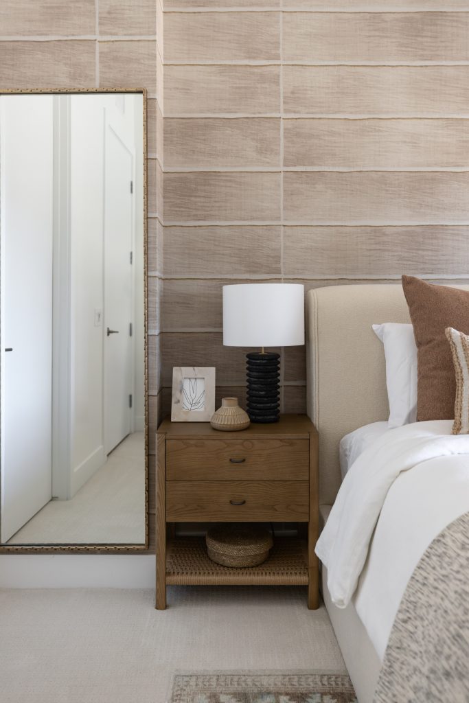 A bedside table with a lamp, framed art, and decor stands next to a bed with neutral bedding; a large mirror rests against the wall.— Living with Lolo.