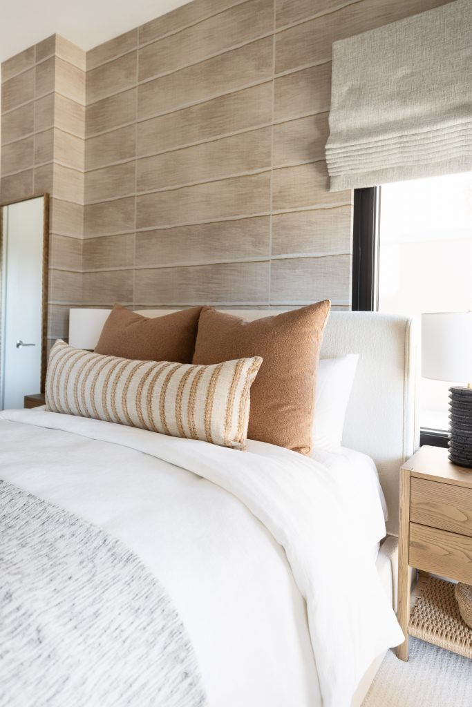 Modern bedroom with a white bed, beige and tan pillows, a wooden nightstand, and a large tiled accent wall behind the bed—Living with Lolo.