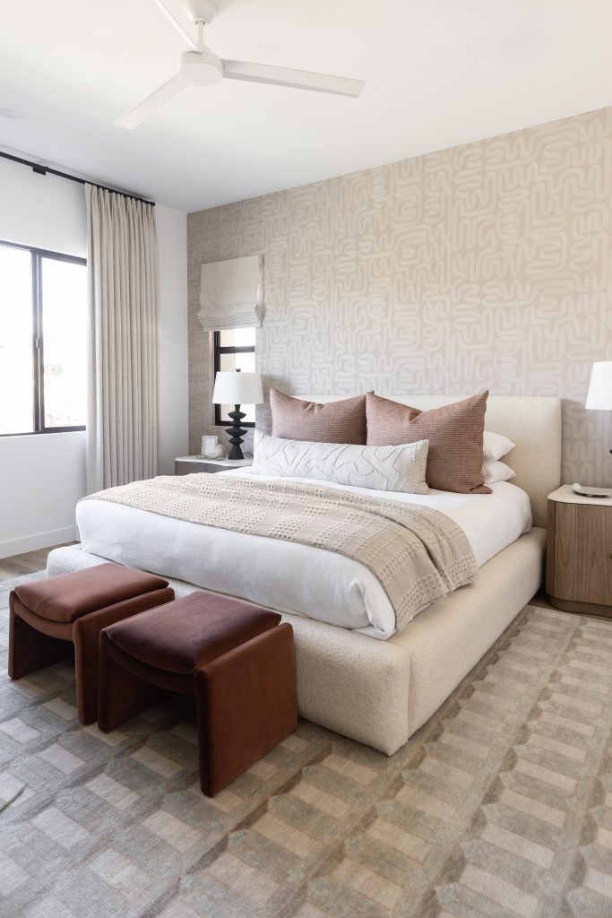 A large upholstered bed with neutral bedding, two brown stools, a geometric rug, and a patterned accent wall by a curtained window — Living with Lolo.