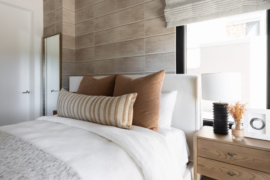 A bed with white linens and brown and striped pillows sits next to a wooden nightstand holding a lamp, photo frame, and dried flowers—Living with Lolo.