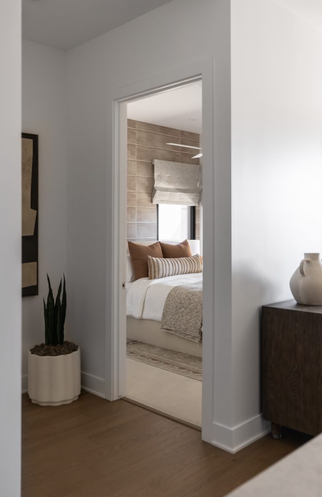 A modern bedroom with neutral colors, a made bed with striped pillows and throw, a plant and vase in front, and a roman shade—Living with Lolo.