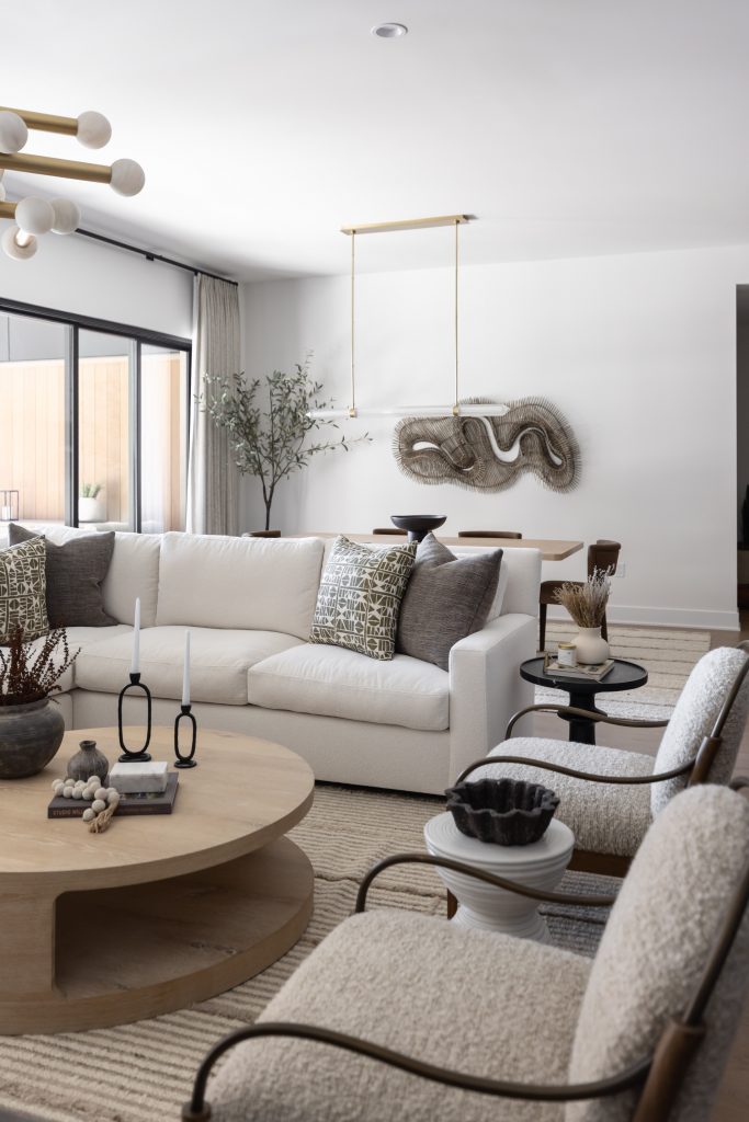 Modern living room with white sofa, round wooden coffee table, armchairs, abstract wall art, and large window. Dining area in back—Living with Lolo.