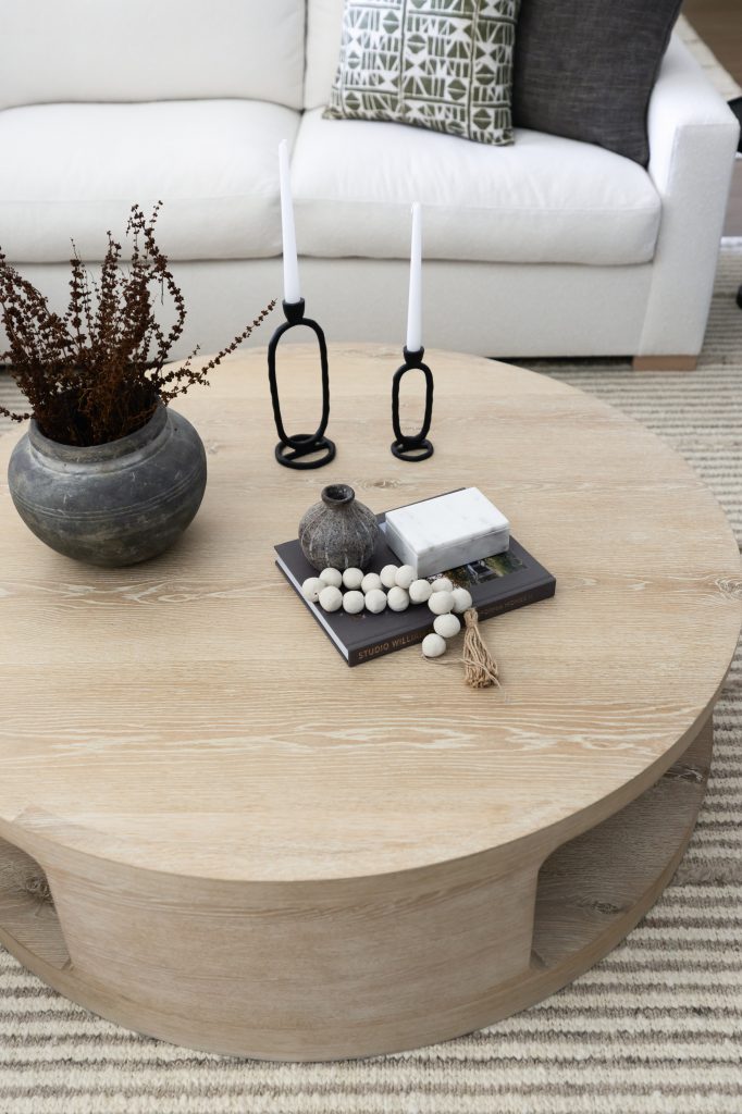A round wooden coffee table holds a vase with dried plants, black candle holders, books, beads, and a ceramic jar—Living with Lolo.