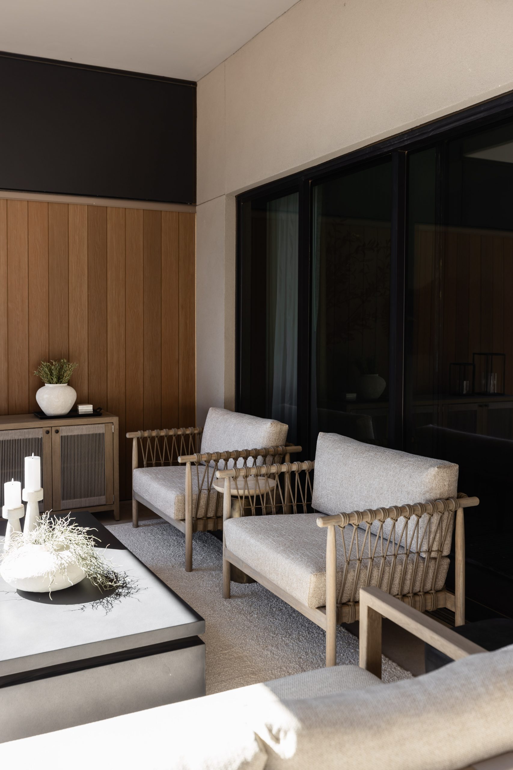 A modern patio with two beige armchairs, a black-framed glass door, a concrete coffee table, and wood-paneled walls. — Living with Lolo.