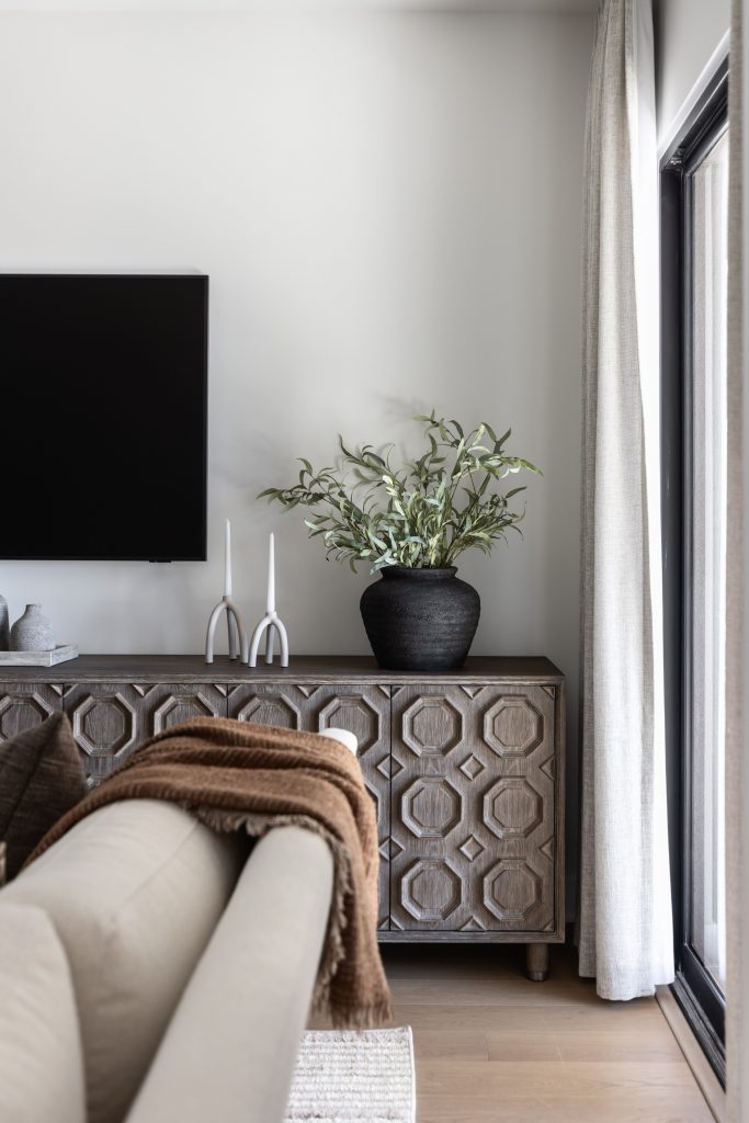 A modern living room with a beige sofa, geometric wooden cabinet, black potted plant, white candle holders, and wall-mounted TV. — Living with Lolo.