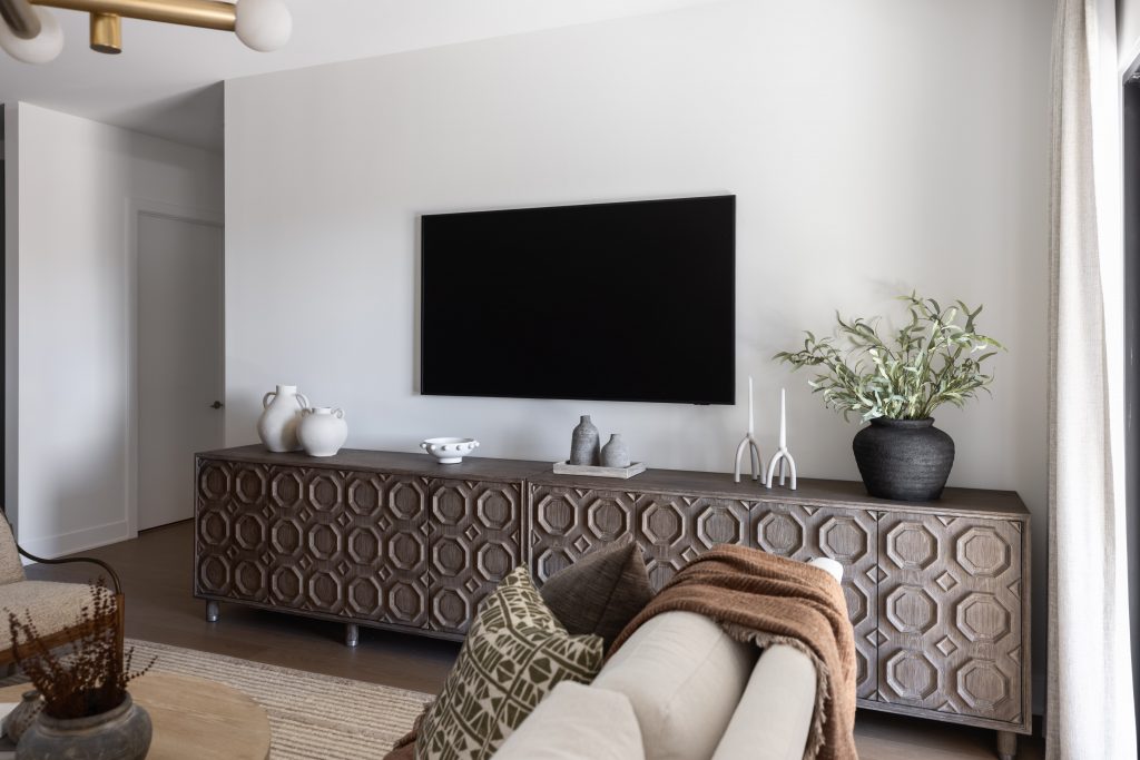 A wall-mounted flat screen TV hangs above a geometric console holding vases and candlesticks, with a beige sofa in front.— Living with Lolo.