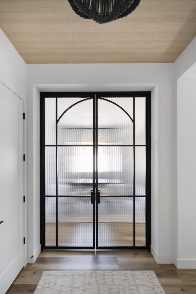 Black-framed double glass doors with arched detailing open to an entryway with white walls, light wood floors, and a rug — Living with Lolo.