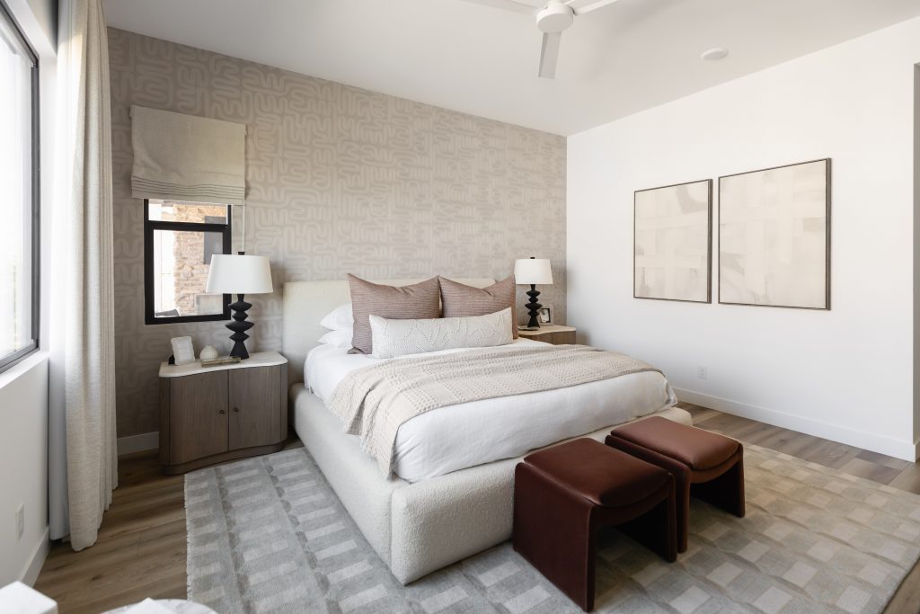 A renovated bedroom with a large bed, beige and brown bedding, bedside tables with lamps, patterned wallpaper, and wall art—Living with Lolo.