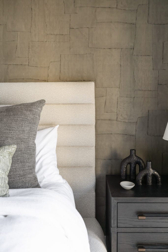 A modern bedroom features a textured beige headboard, gray pillows, a dark nightstand with sculptural vases, and a stone-patterned wall—Living with Lolo.
