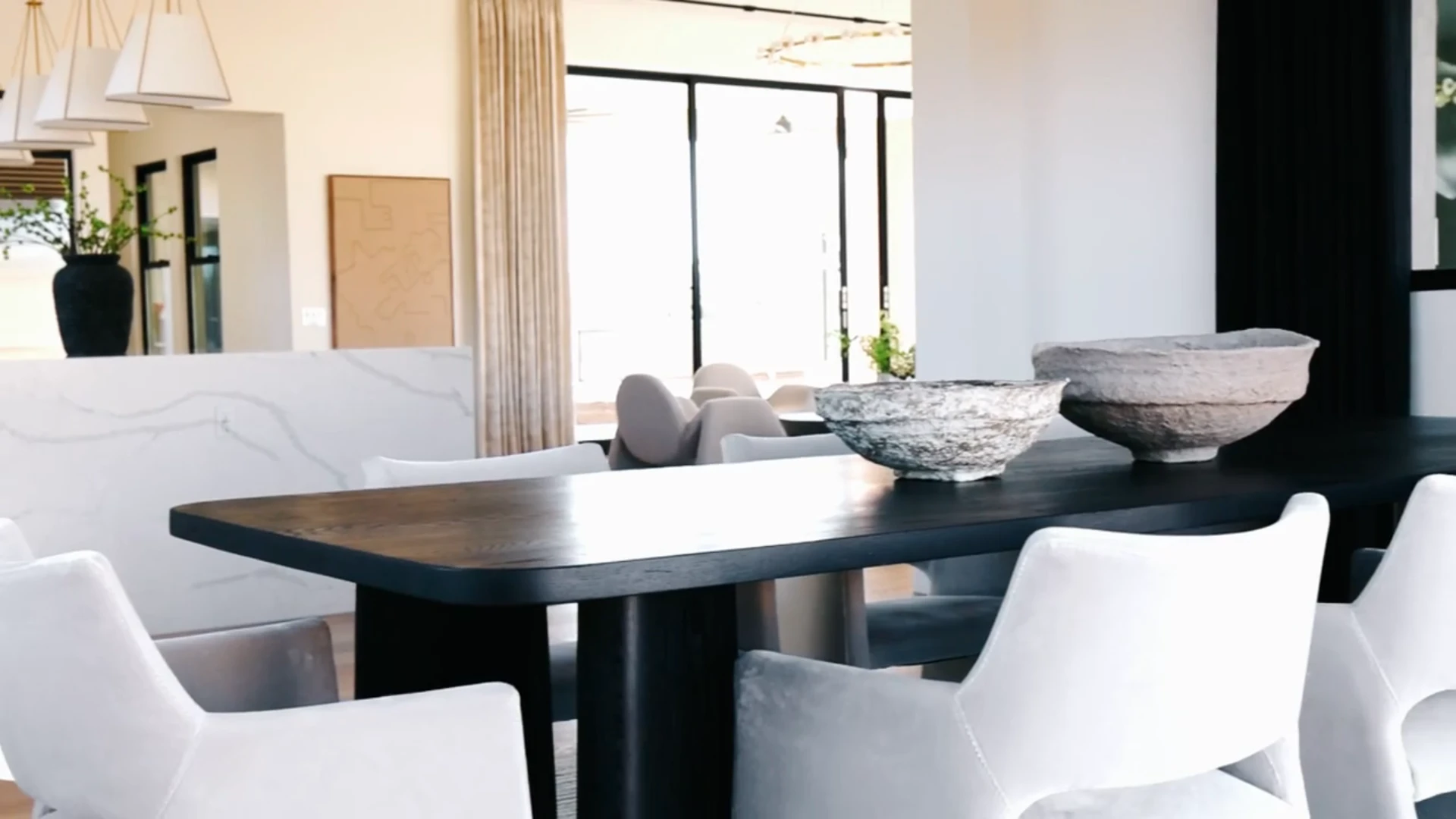 A dark rectangular dining table, white upholstered chairs, stone bowls, and large windows define the neutral-toned space. — Living with Lolo. A dark rectangular dining table, white upholstered chairs, stone bowls, and large windows define the neutral-toned space. — Living with Lolo.