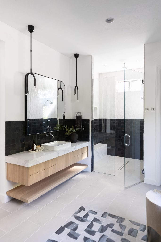 Modern bathroom with a floating wood vanity, large mirror, black tile accent wall, glass shower enclosure, and a geometric-patterned rug on a light tile floor—Living with Lolo.