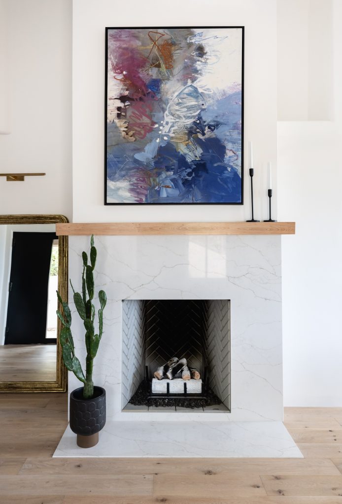 A modern white fireplace features a potted cactus, two tall candlesticks, and an abstract painting above the mantel. A large mirror leans against the wall to the left — Living with Lolo.