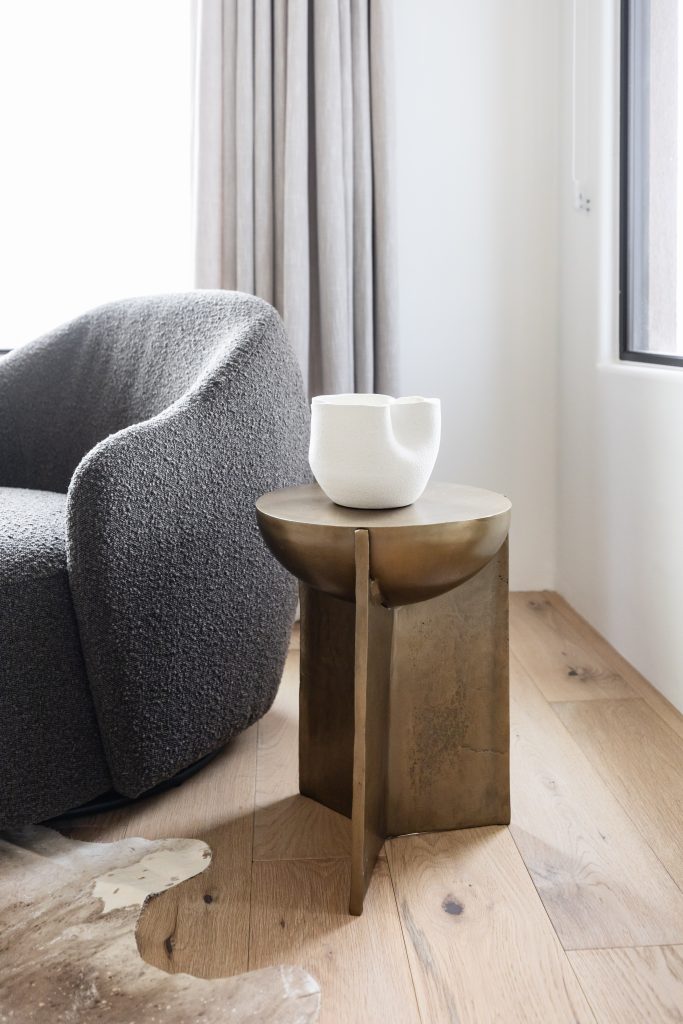 A gray textured chair is positioned beside a metallic side table holding a white ceramic bowl, set on light wood flooring next to a window and beige curtain—Living with Lolo.