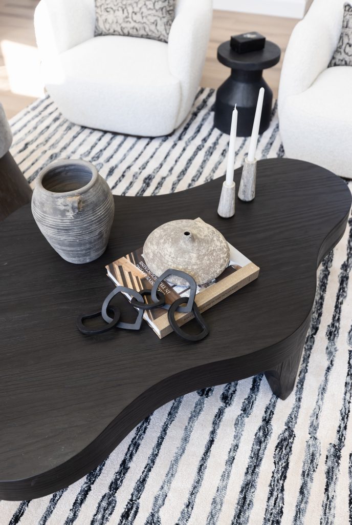 Black coffee table with ceramic vases, two white candles, books, and decorative objects on a striped rug, with white armchairs in the background.— Living with Lolo.