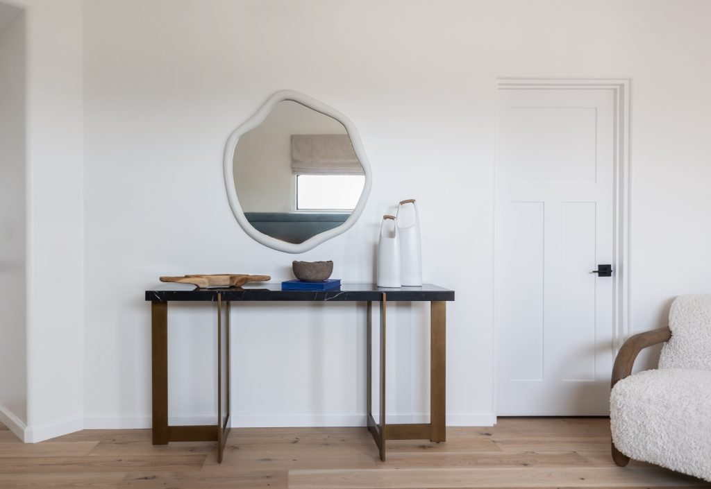 A black console table with gold legs stands beneath a round mirror in an entryway. A closed white door and part of a textured armchair are also visible — Living with Lolo.