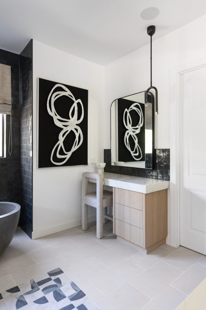 A modern bathroom features a light wood vanity, black sink backsplash, abstract black-and-white wall art, a circular chair, and a white tile floor with a geometric rug. — Living with Lolo.