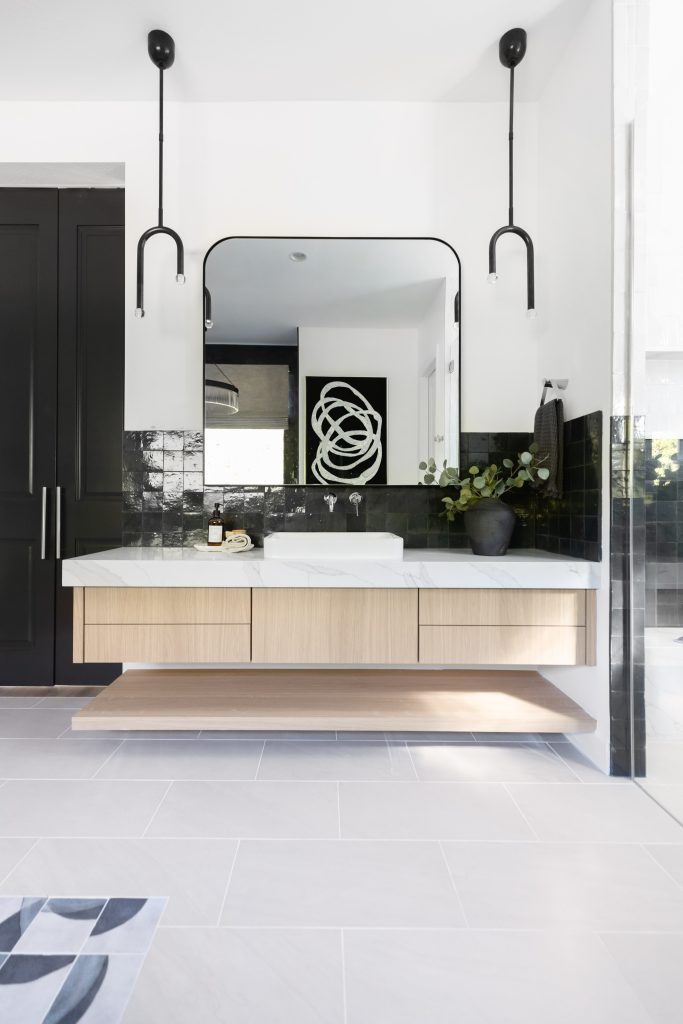 Modern bathroom featuring a floating wood vanity, rectangular mirror, black tile backsplash, two pendant lights, and a black vase with greenery on the counter—Living with Lolo.