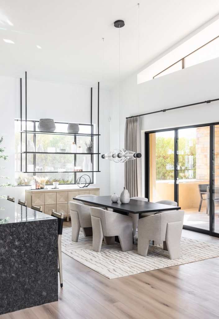 Modern dining room featuring a black table, six upholstered chairs, pendant lights, large windows, sliding glass doors, and a sideboard with decorative items—Living with Lolo.