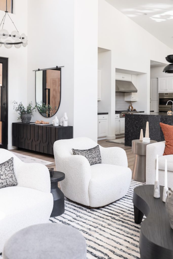 Modern living room featuring white boucle chairs, a striped rug, and black accent tables. The space opens to a kitchen with white cabinets and a dark island—Living with Lolo.