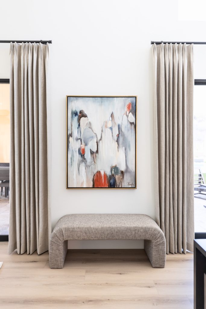 A modern bench is placed beneath an abstract painting between two beige curtains in a bright room with light wood flooring—Living with Lolo.