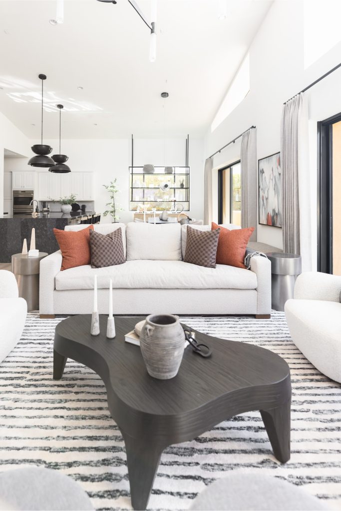 A neutral sofa with orange accent pillows sits next to an abstract coffee table and a striped rug. The background shows a minimalist kitchen with pendant lighting—Living with Lolo.