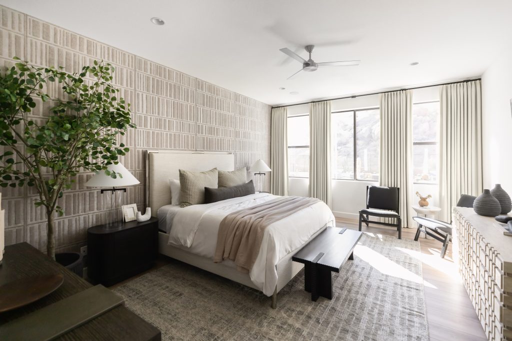 Modern bedroom featuring a large bed with neutral bedding, a bench at the foot, patterned wallpaper, an indoor plant, tall windows providing natural light, and minimalist decor—Living with Lolo.