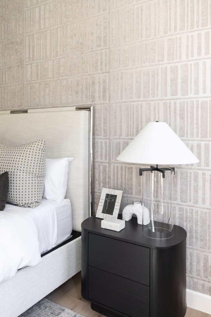 Modern bedroom featuring a white upholstered bed with patterned pillows, a black nightstand holding a lamp and decorative object, and framed art on a textured beige wall—Living with Lolo.