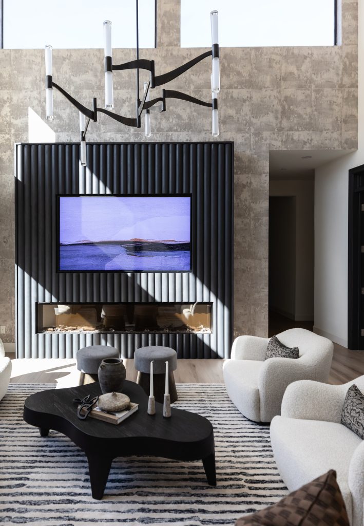 Modern living room with a textured accent wall, wall-mounted TV, black coffee table, white armchairs, patterned rug, and decorative accessories. Natural light enters through high windows—Living with Lolo.