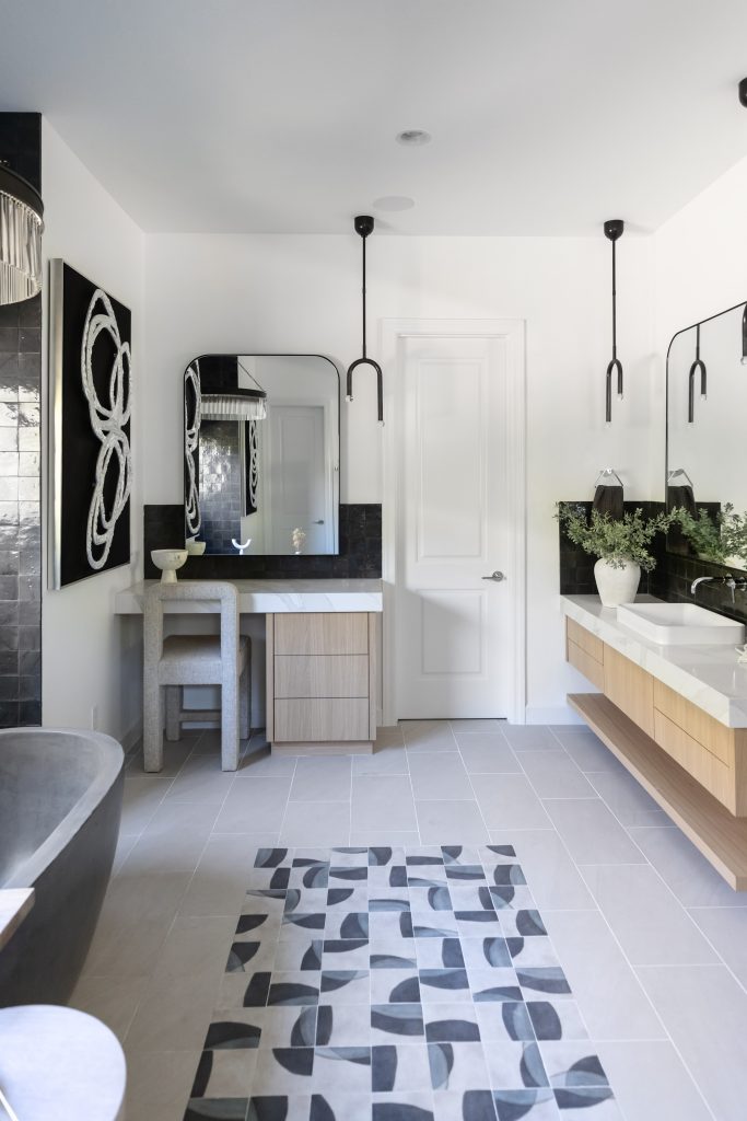 A modern bathroom with a freestanding tub, double sinks, black and white decor, a geometric rug, light wood cabinetry, and wall-mounted mirrors with black sconces. — Living with Lolo.