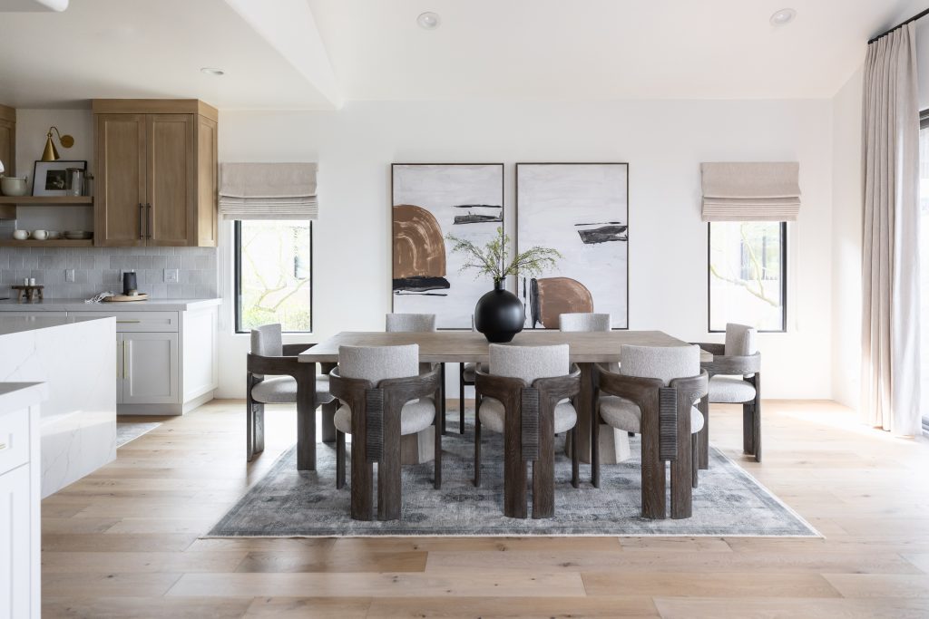A dining room features a dark wood table surrounded by eight upholstered chairs, light wood floors, two abstract art pieces on the wall, and a black vase with greenery on the table. Natural light fills the space — Living with Lolo.