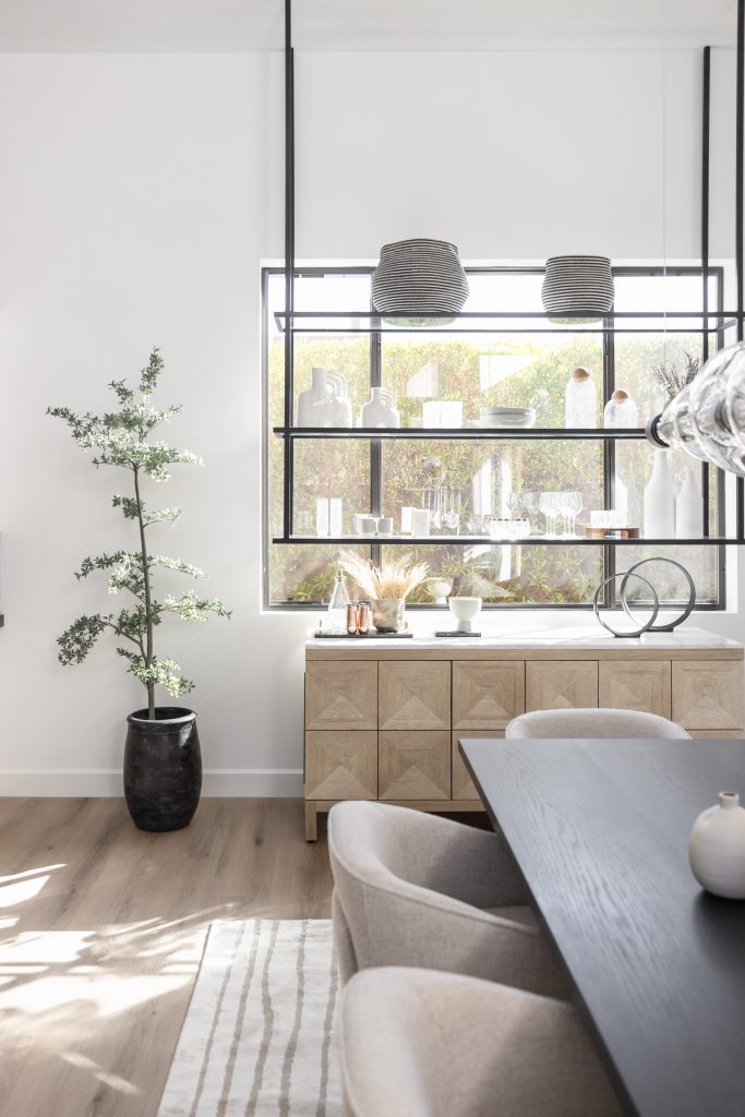A dining room with a black table, beige chairs, a potted plant, and shelves of decorative items is set in front of a large window. — Living with Lolo.