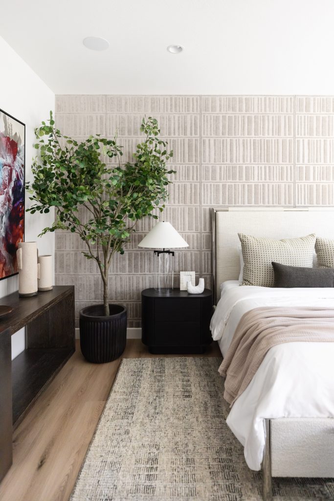 Modern bedroom with neutral tones, a large potted plant beside the bed, artwork on the wall, a nightstand with a lamp, and a neatly made bed with pillows and a beige throw blanket. — Living with Lolo.