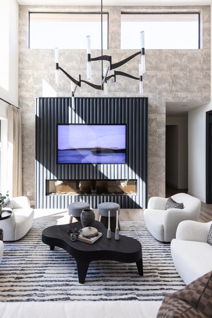 Modern living room featuring a large TV, black abstract coffee table, white armchairs, a striped rug, and geometric chandelier below two tall windows—Living with Lolo.