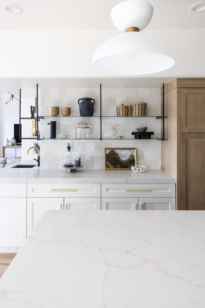 Modern kitchen featuring white marble countertops, open glass shelves with pottery and decor, white cabinets, brass hardware, and a central overhead light fixture—Living with Lolo.