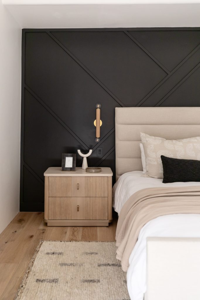 A bedroom with a black accent wall, beige headboard, light nightstand, wall sconce, neutral bedding on a made bed, wood floor, and patterned rug is shown. — Living with Lolo.