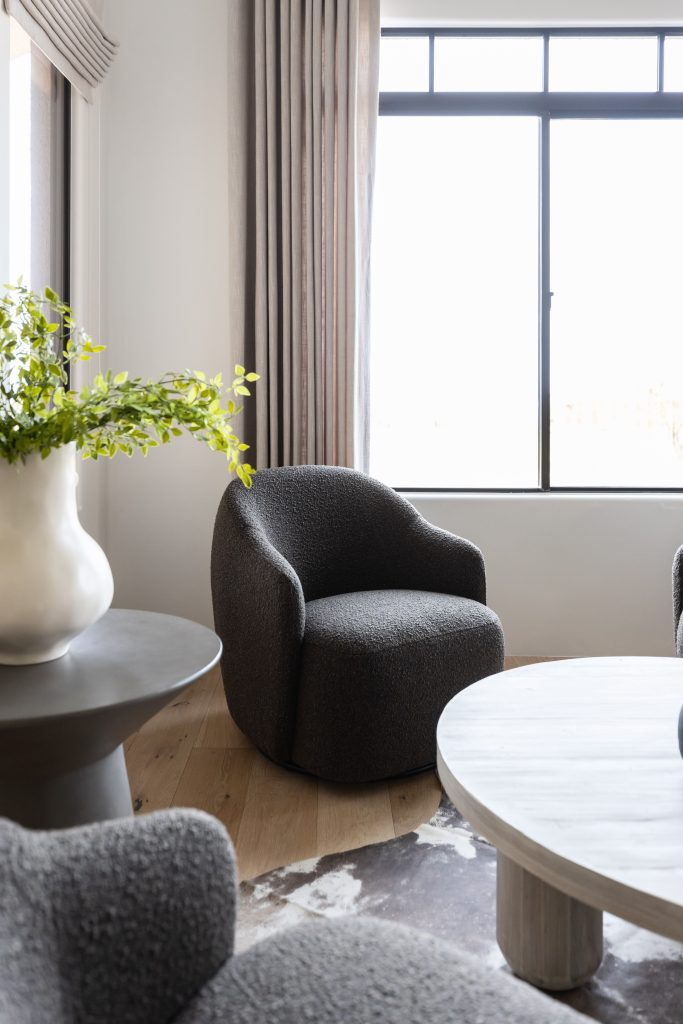 A modern living room features a textured gray armchair, a round coffee table, a side table holding a white vase with greenery, and a large window with light curtains. — Living with Lolo.