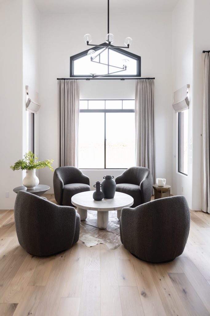 Modern living room featuring four dark armchairs arranged around a white round coffee table, a large window with light curtains, and a contemporary chandelier overhead—Living with Lolo.