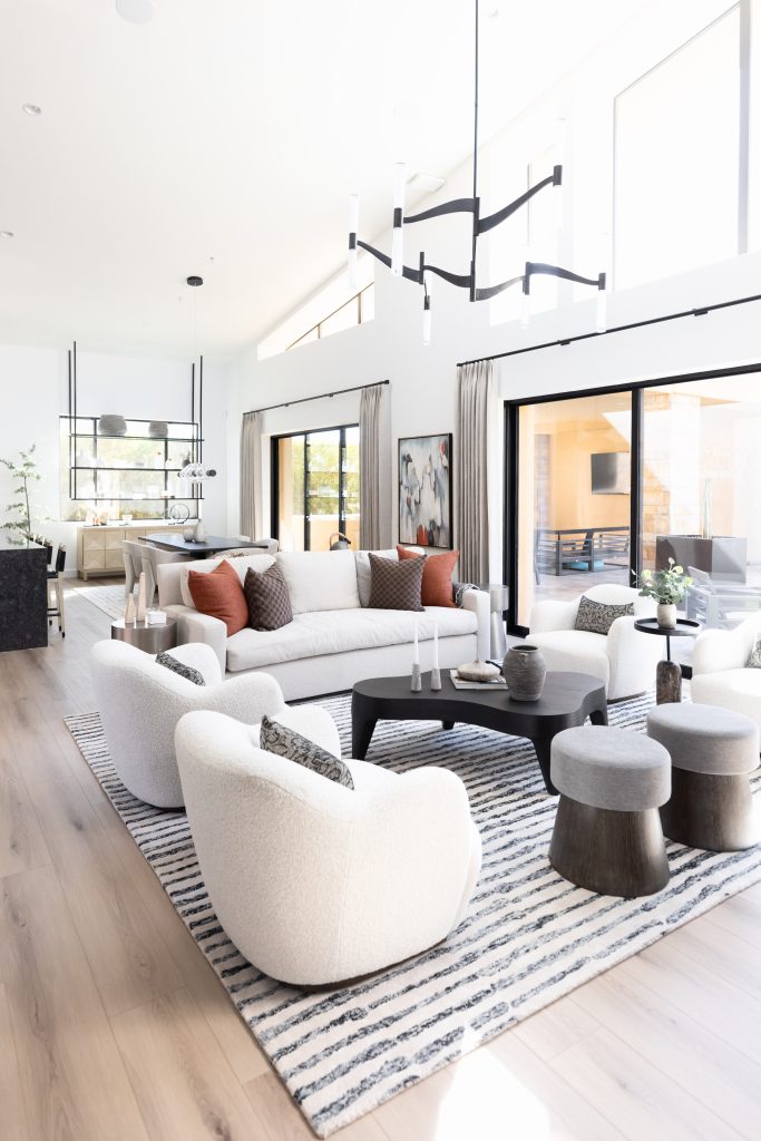 Modern living room featuring white curved chairs, a gray sofa, black coffee tables, a striped rug, and large windows. A dining area is visible in the background—Living with Lolo.