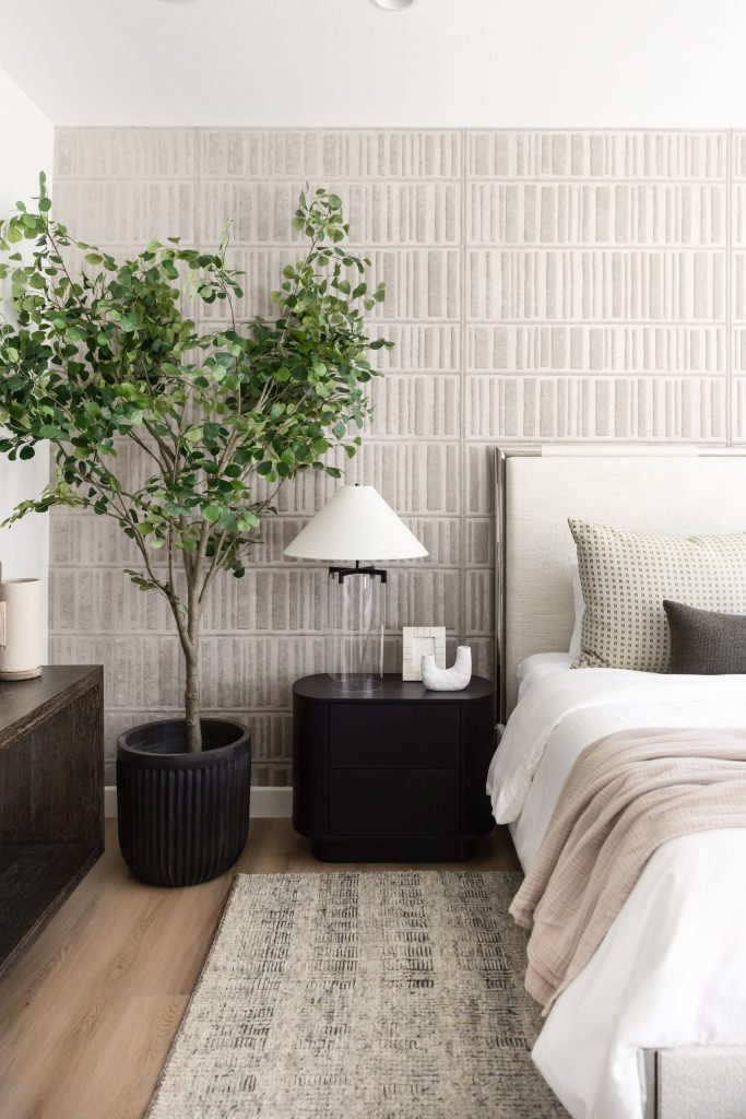 A bedroom with a potted tree, black nightstand, lamp, decorative object, beige upholstered bed, and patterned wallpaper is shown. — Living with Lolo.