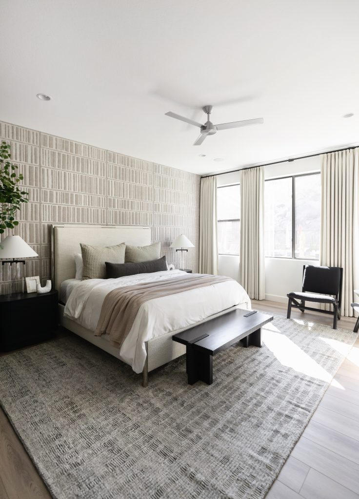 Modern bedroom featuring a large bed with neutral bedding, two black nightstands, a black bench, a black chair, light patterned wallpaper, large windows with curtains, and a ceiling fan—Living with Lolo.